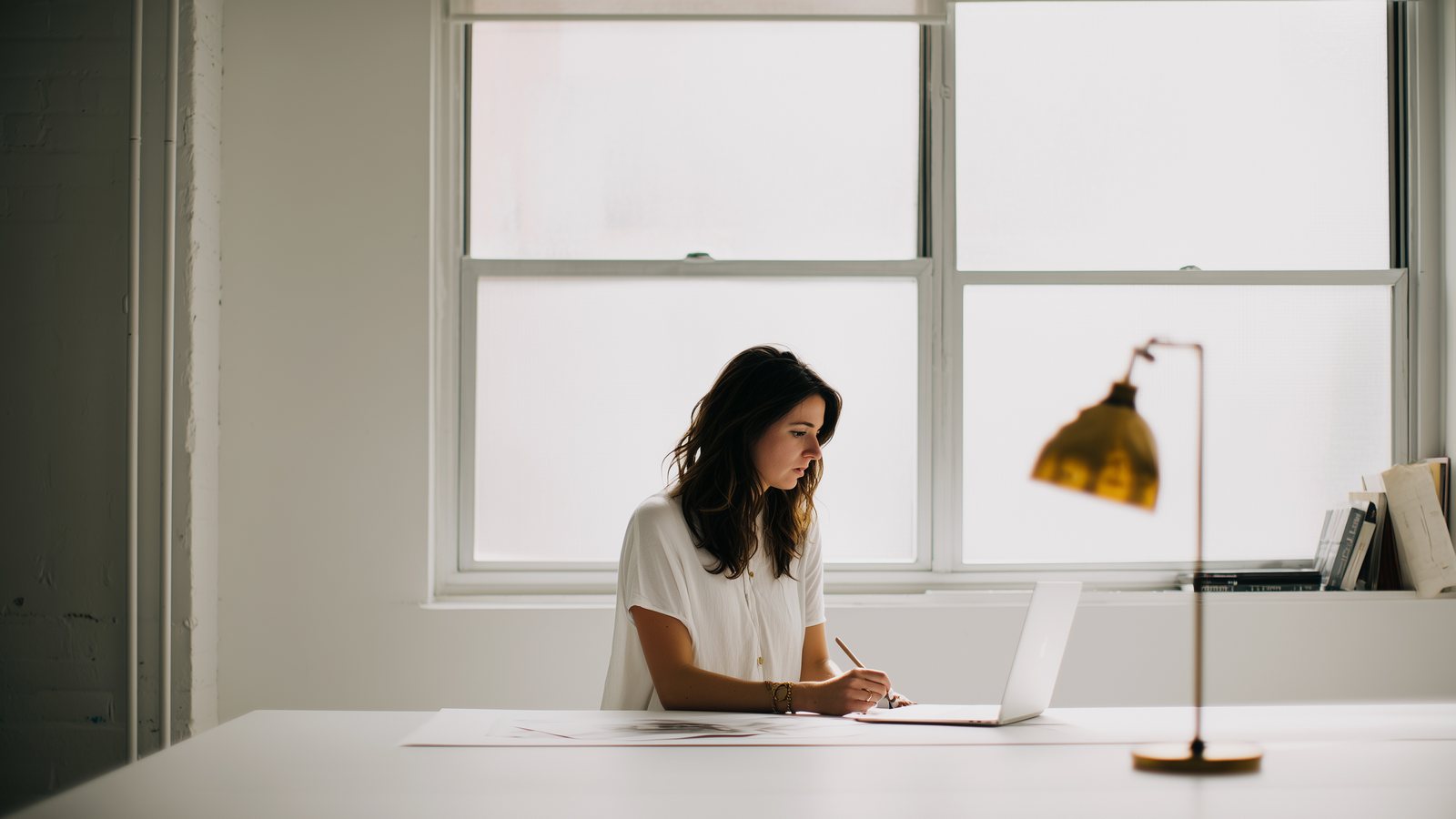 Someone working alone at a desk by a window, laptop open and notebook beside it
