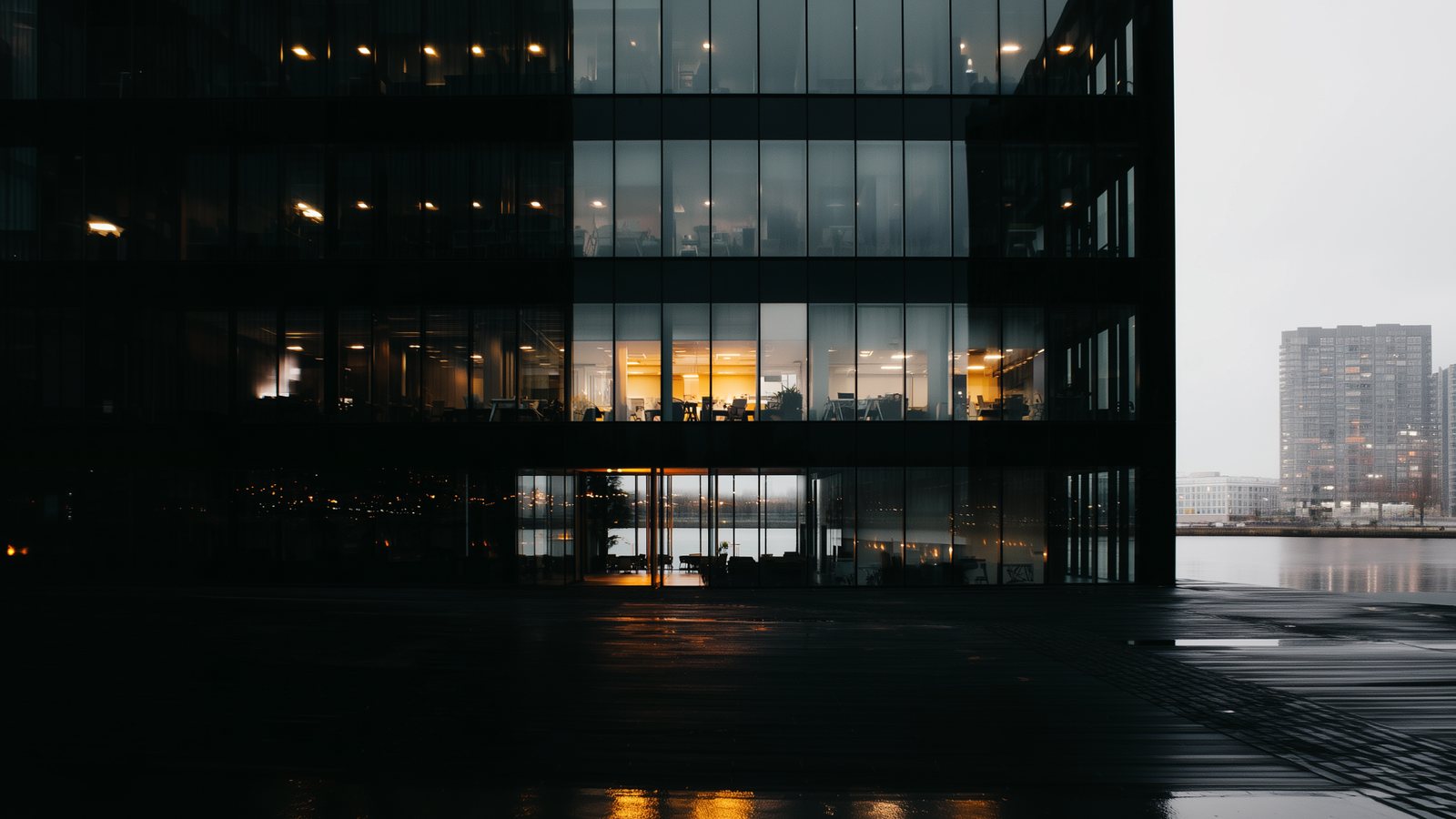 A dark office building at night beside water, amber light reflected on the quay